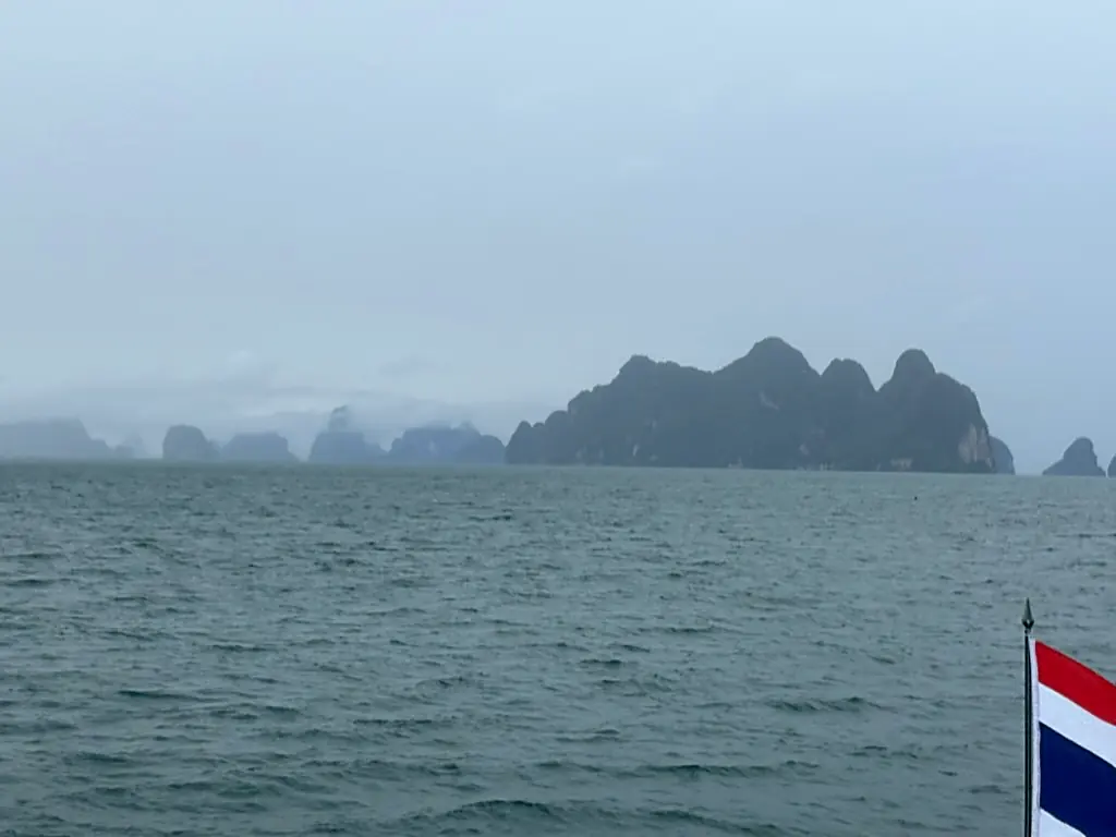 Limestone islands fading into grey mist across Phang Nga Bay, a Thai flag at the stern