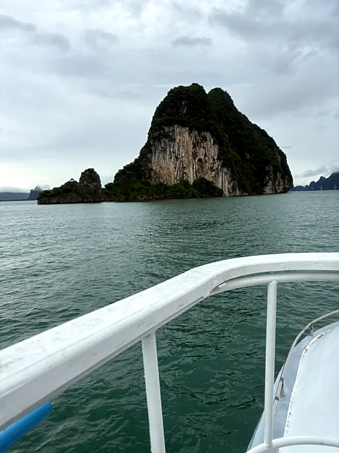 A single forested limestone karst seen over the white bow rail of a boat